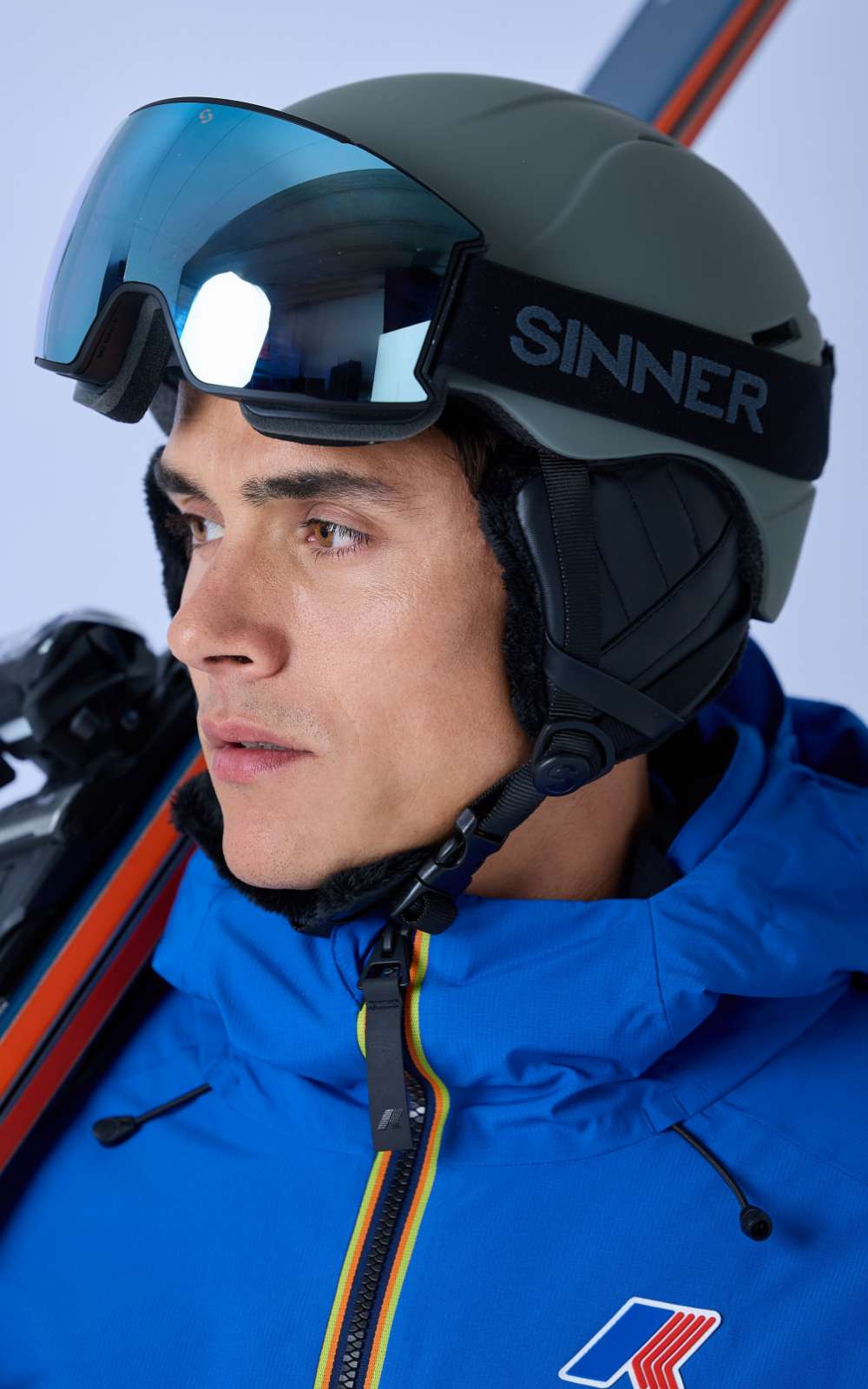 Skiër met helm en grote skibril kijkt opzij terwijl ski’s achter de schouder zichtbaar zijn.