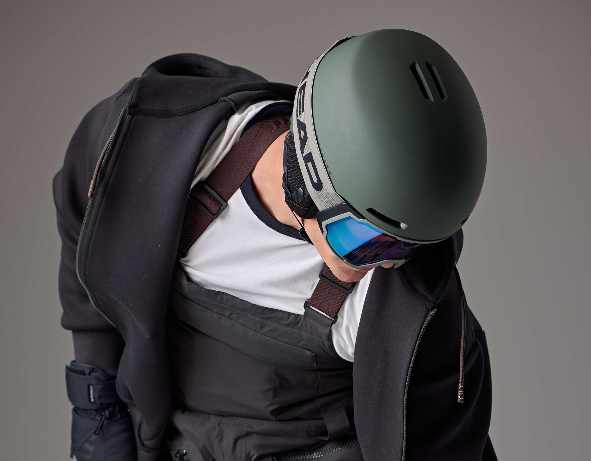 Persoon in ski-outfit draagt een beige skihelm met skibril met opschrift “APEX”, zichtbaar van opzij tegen een neutrale achtergrond.