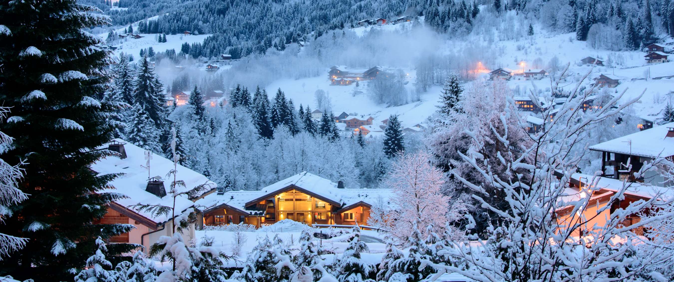 Besneeuwde chalets tussen winterbomen met warme avondverlichting.