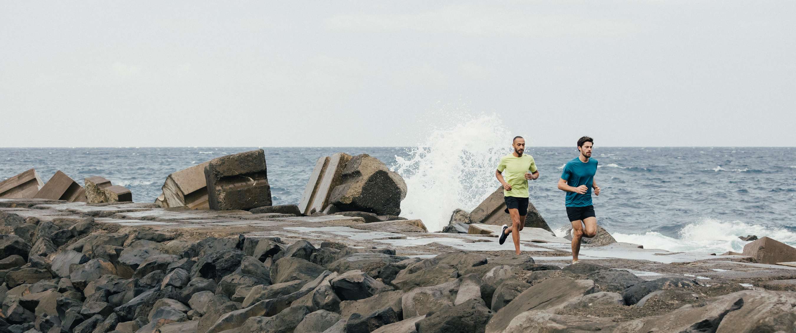 Twee hardlopers lopen langs rotsen bij zee met opspattende golven.