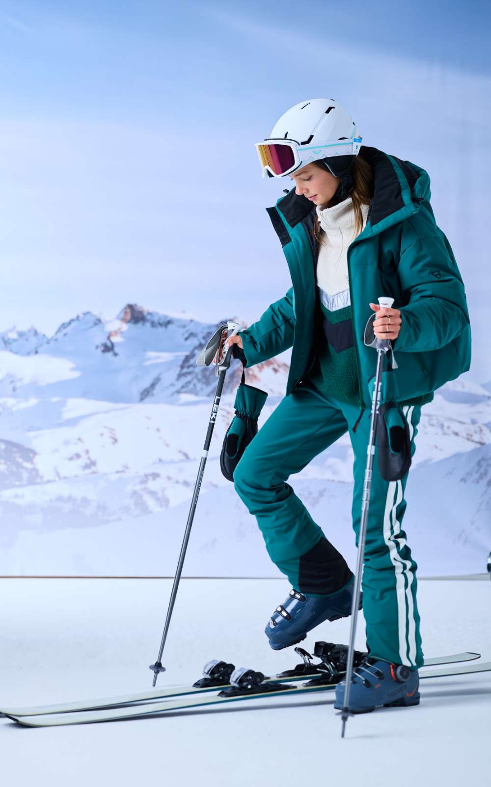 Een skiër met witte helm en skibril kijkt omlaag naar haar ski's tegen een achtergrond met sneeuwbergen.