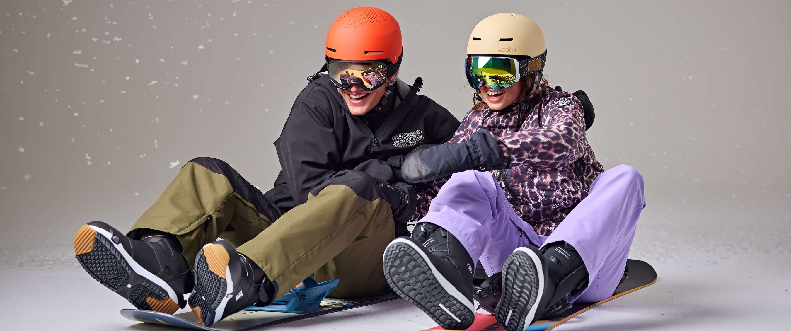 Twee snowboarders die zittend lachen in studio-opstelling met sneeuw-effect.