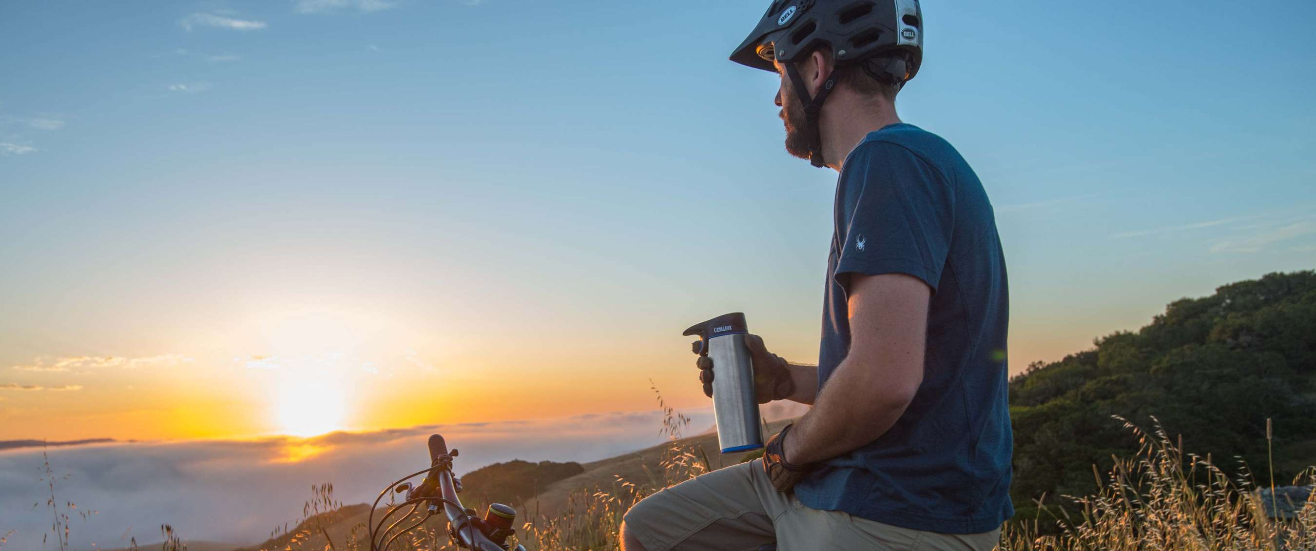 Fietser met helm zit bij een mountainbike en houdt een metalen drinkfles vast bij zonsondergang.